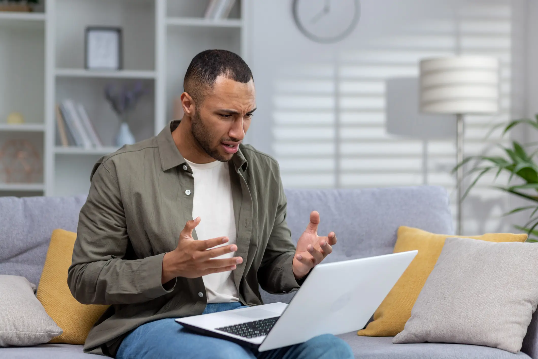 Worried young African-American man at home on sofa holding laptop and talking on video call with psychologist, undergoing online therapy and consultation.